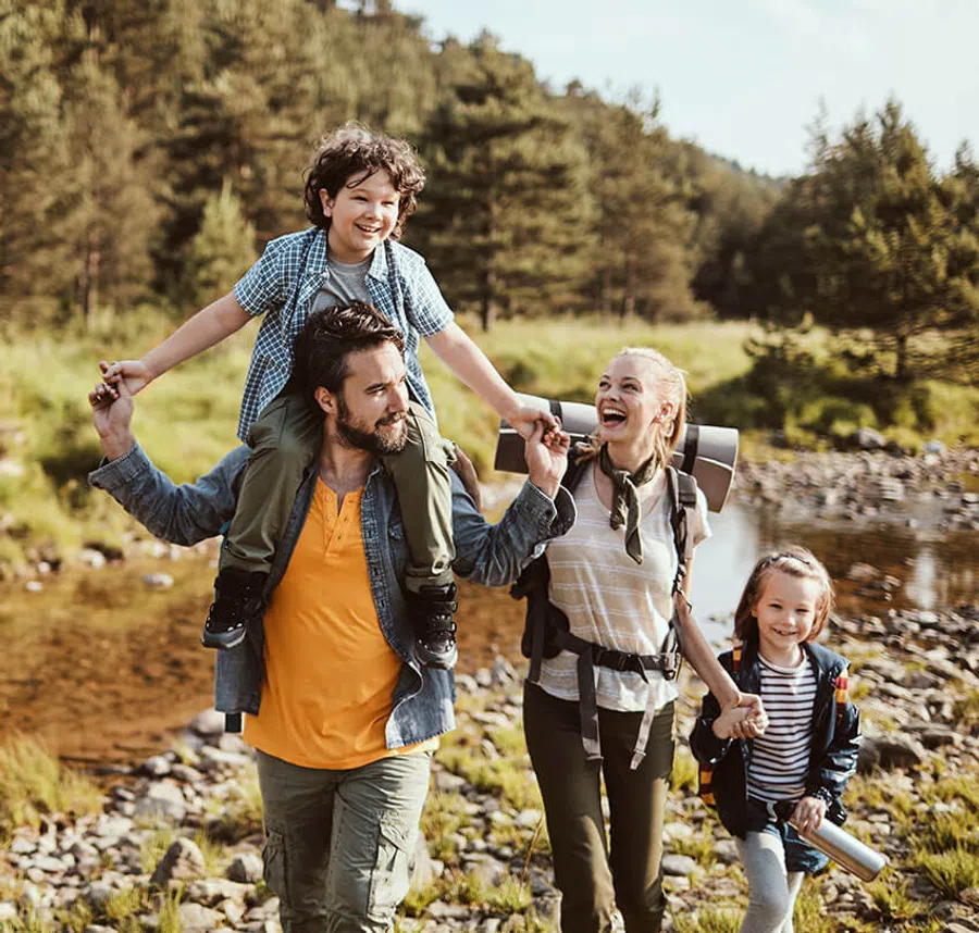 Happy family hiking in nature by a river, father carrying son on shoulders, mother and daughter holding hands.