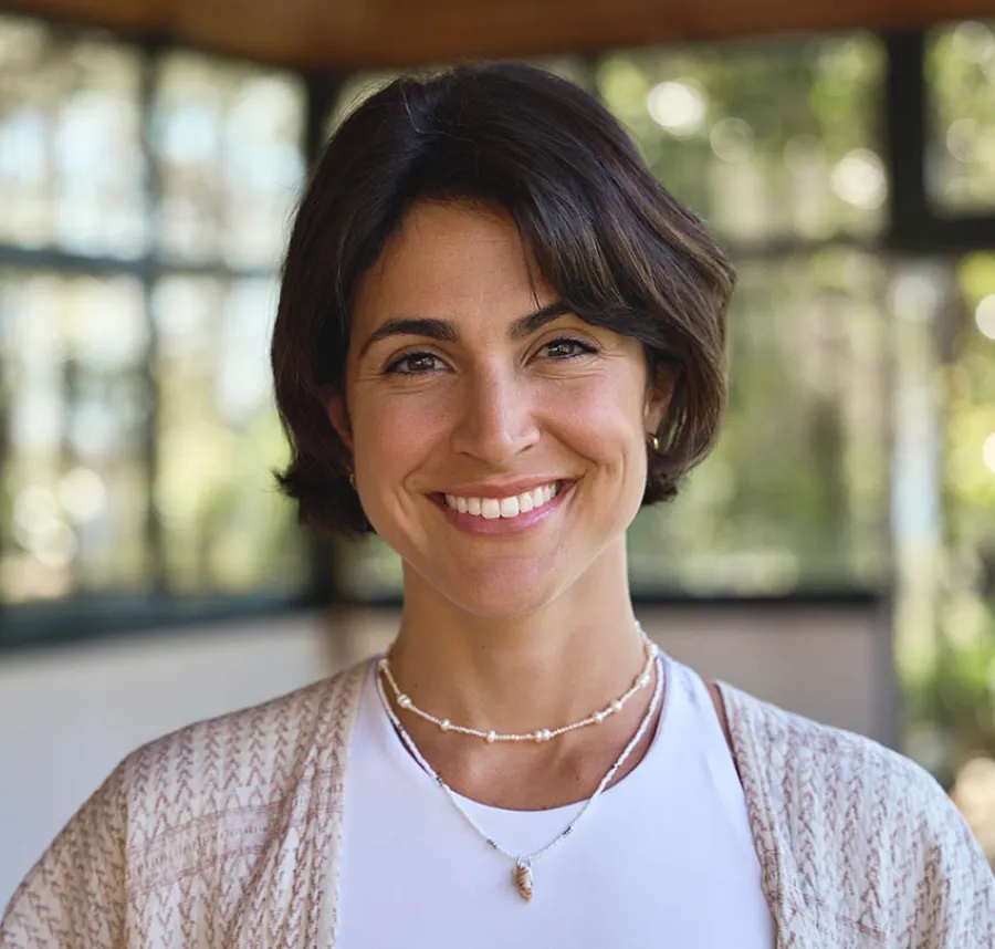 Smiling woman with short dark hair wearing layered necklaces and a beige cardigan indoors with blurred background