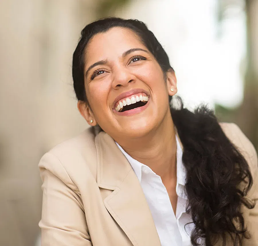 Close-up of a joyful woman with dark hair laughing, wearing a beige blazer and white shirt indoors.
