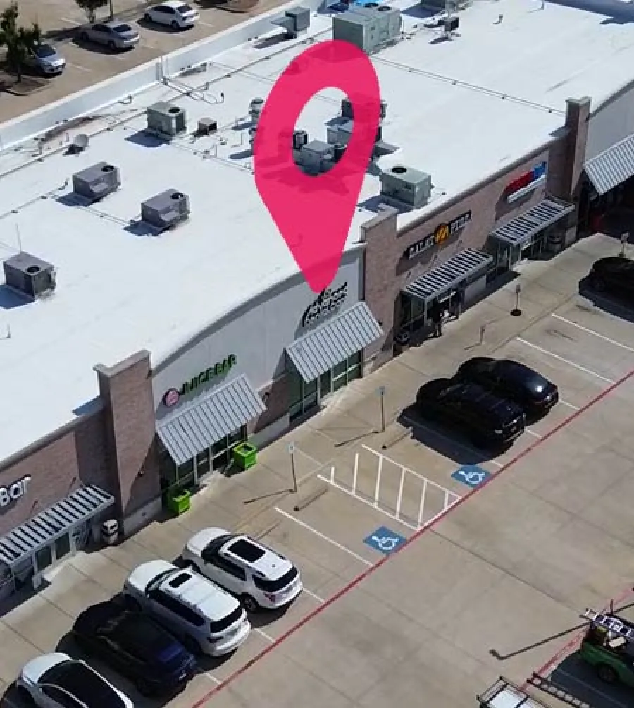 Aerial view of a shopping strip with storefronts, parking spaces, and a red location pin icon above one store.