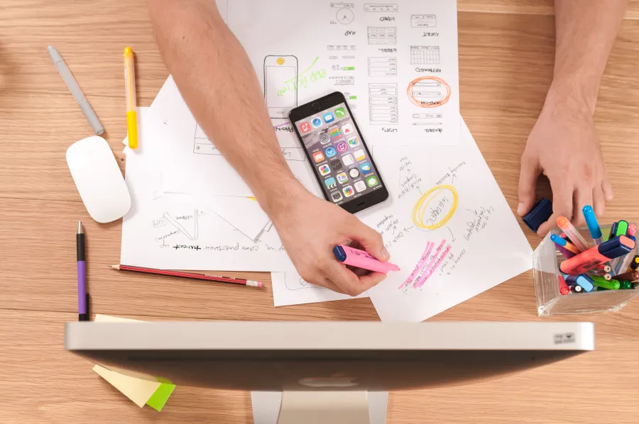 Person highlighting notes on paper near smartphone and colorful markers on wooden desk with computer screen.