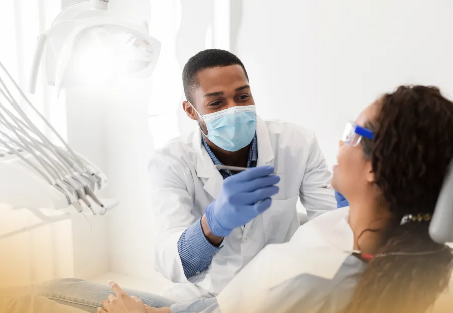 Dentist wearing mask and gloves examining patient in dental clinic with bright light and equipment.