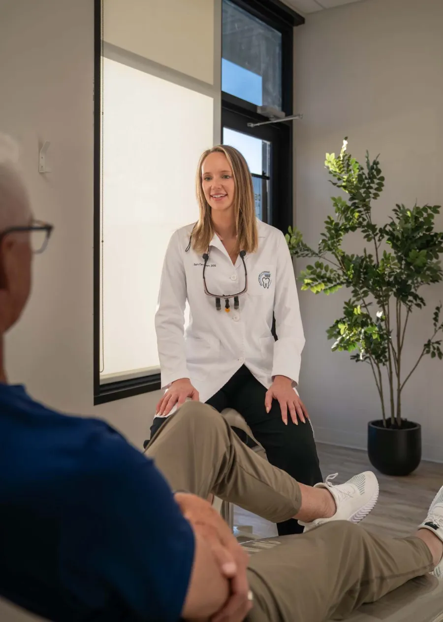 Dentist in white coat talking to patient in a modern dental office with natural light and plants.