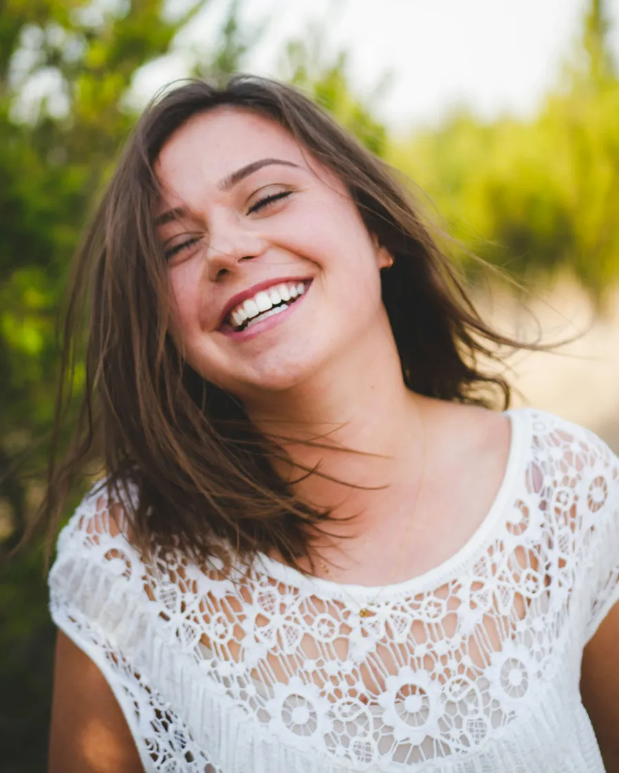 Smiling young woman with brown hair wearing a white lace top in an outdoor natural setting.