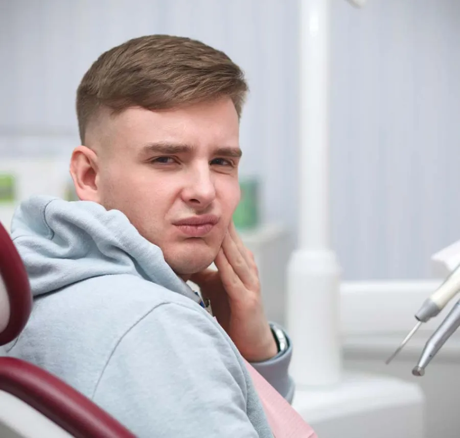 Young man sitting in dental chair with a painful expression, holding his cheek in discomfort.