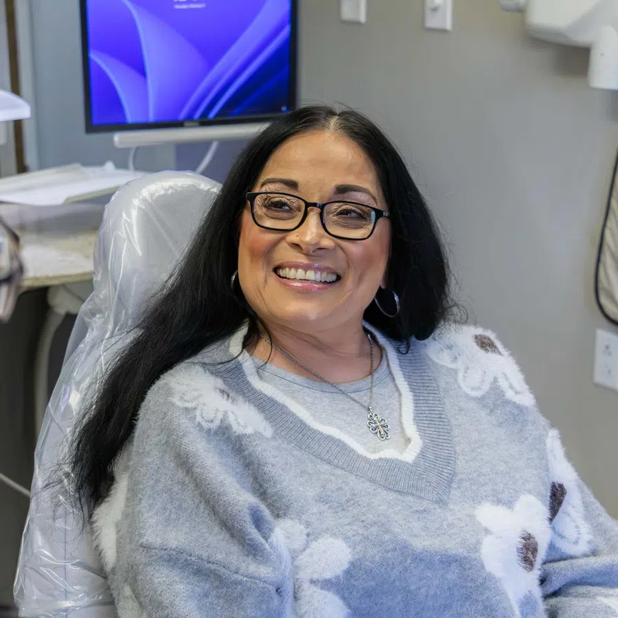 Smiling woman with glasses sitting in dental chair talking to a dental professional with computer screen in background