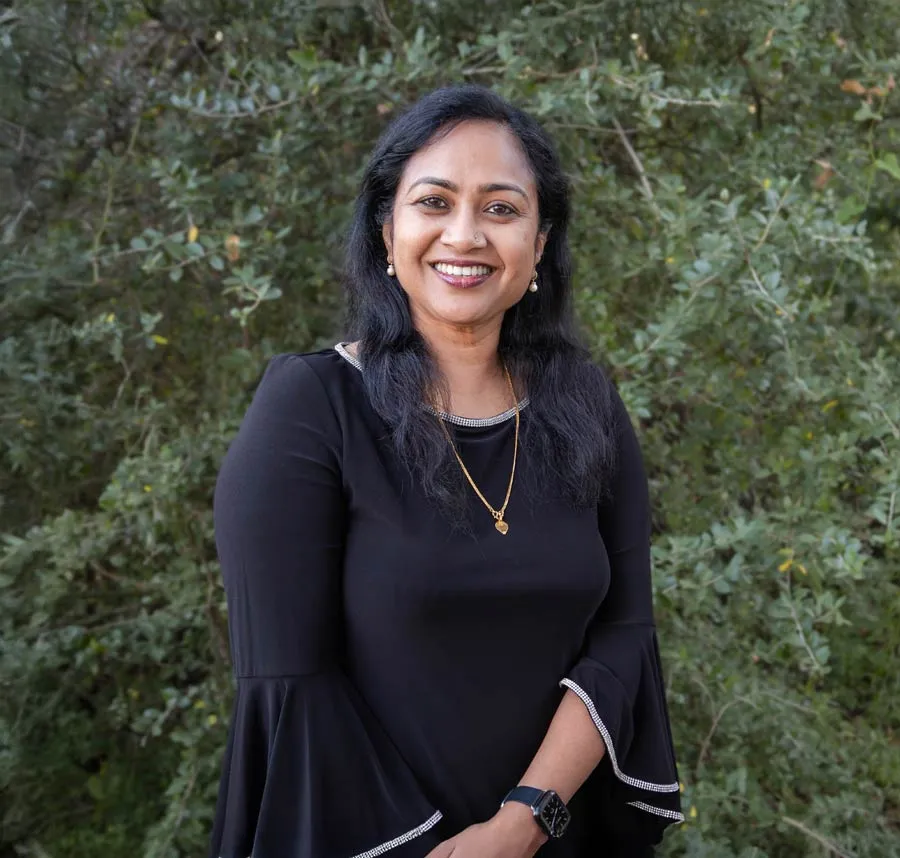Smiling woman in black dress with gold necklace stands outdoors with green foliage background.