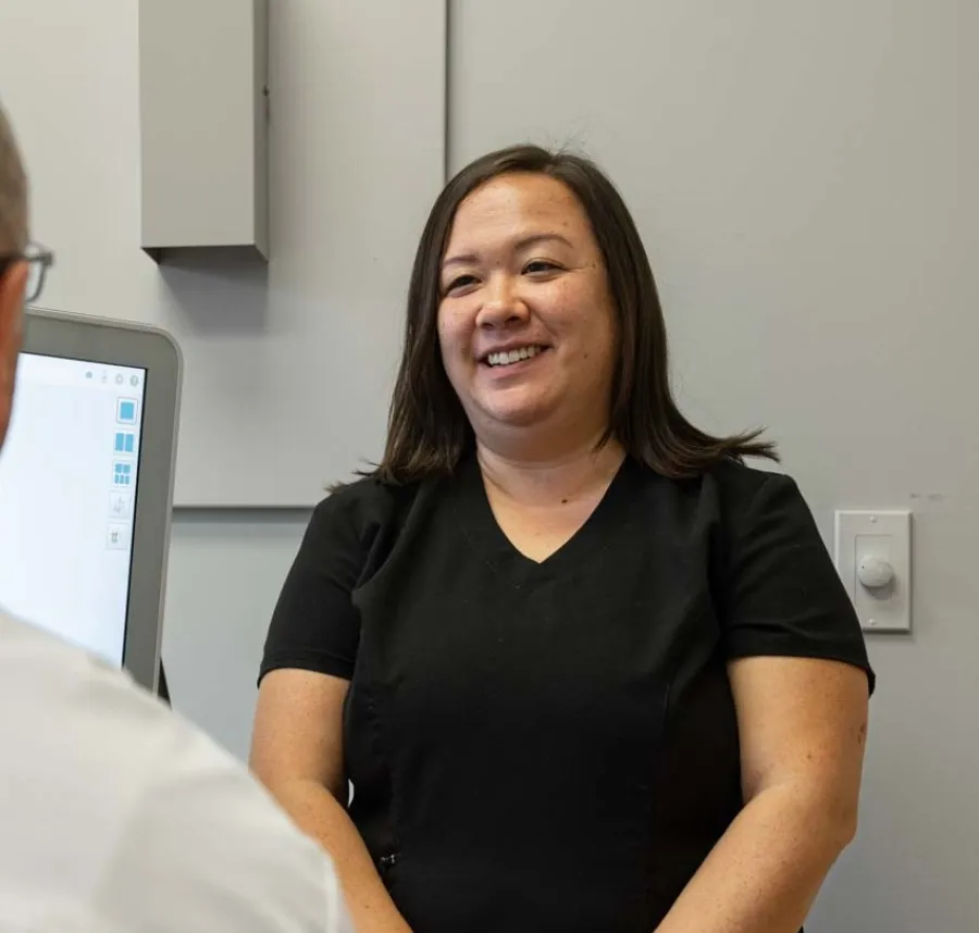 Smiling female healthcare professional talks with senior male patient in medical office with computer screen.