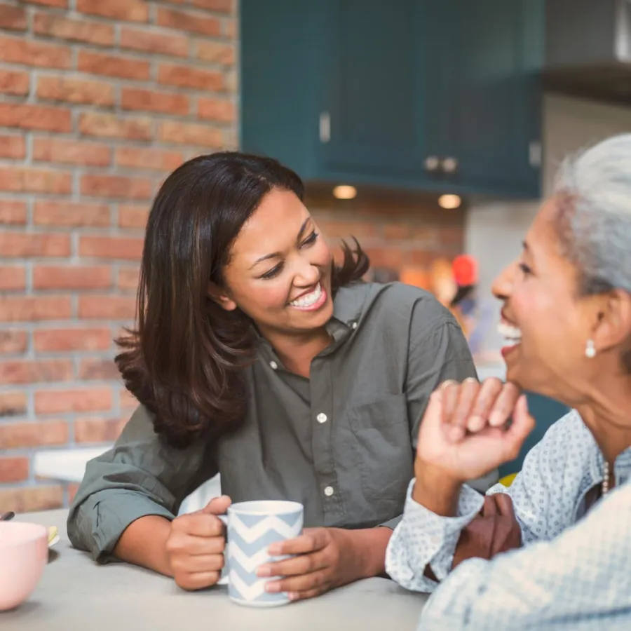 Two women laughing and enjoying coffee together at a kitchen table with a brick wall background.