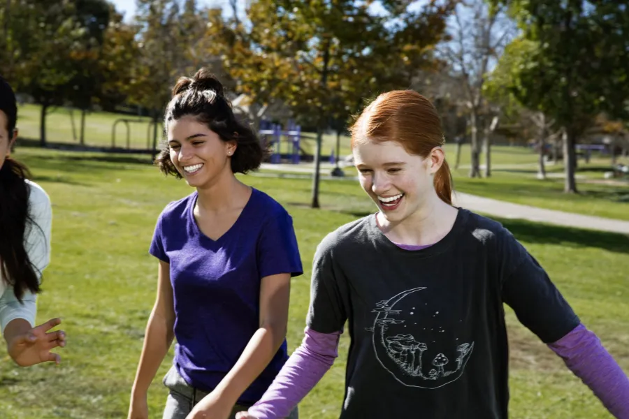 Two smiling teenage girls enjoying a sunny day outdoors in a park with green grass and autumn trees.