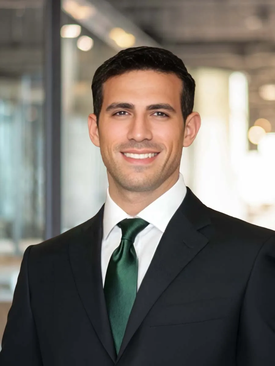 Smiling professional man in black suit and green tie poses in modern office setting with blurred background.