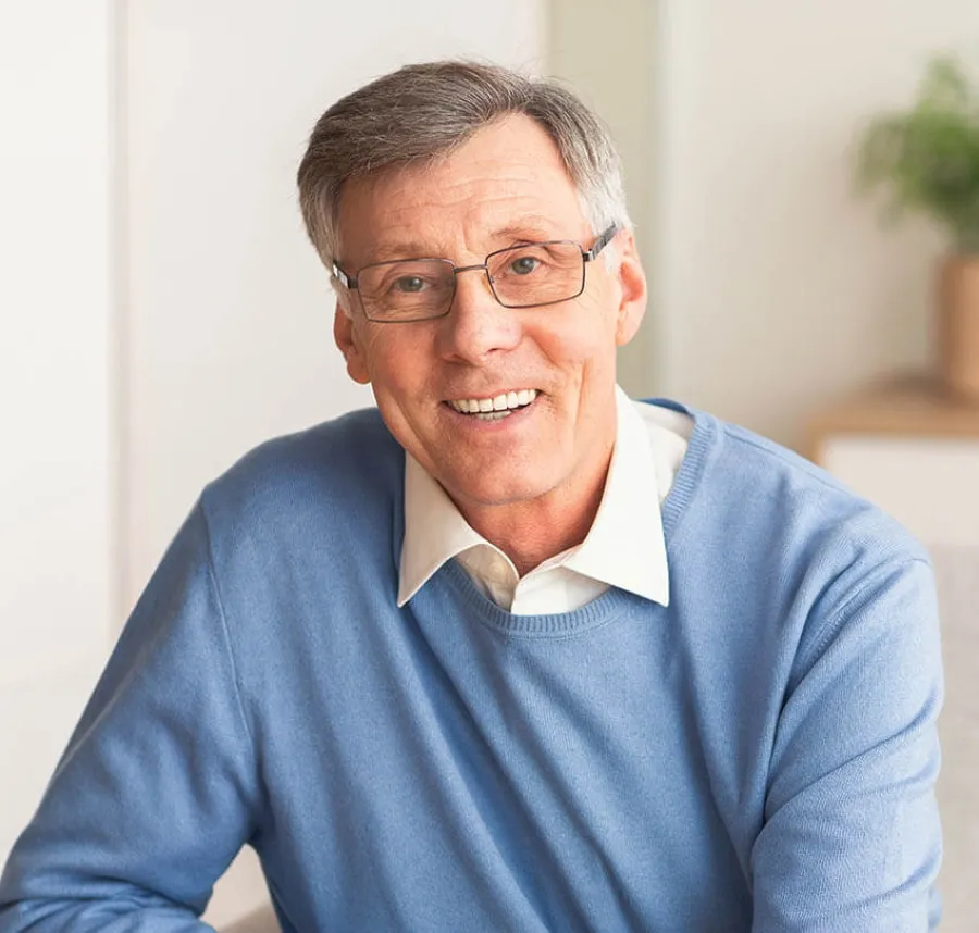 Smiling mature man wearing glasses and blue sweater sitting indoors with soft background and natural light.