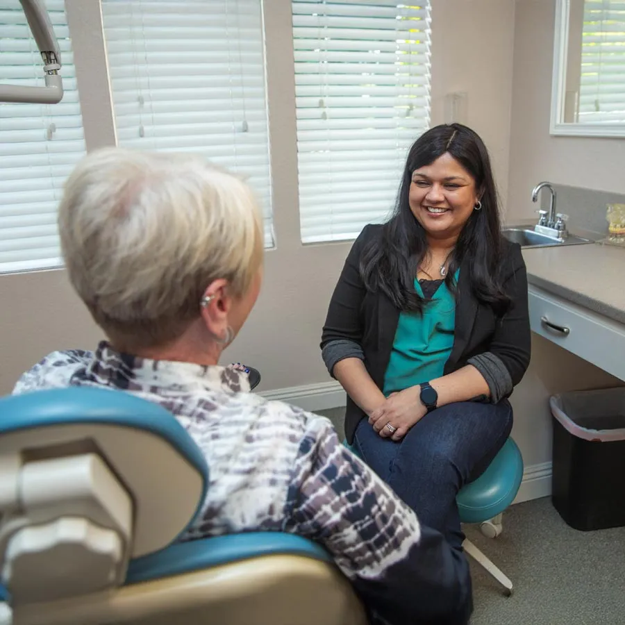 Smiling healthcare professional talking with elderly patient in a bright medical office with blinds and sink.