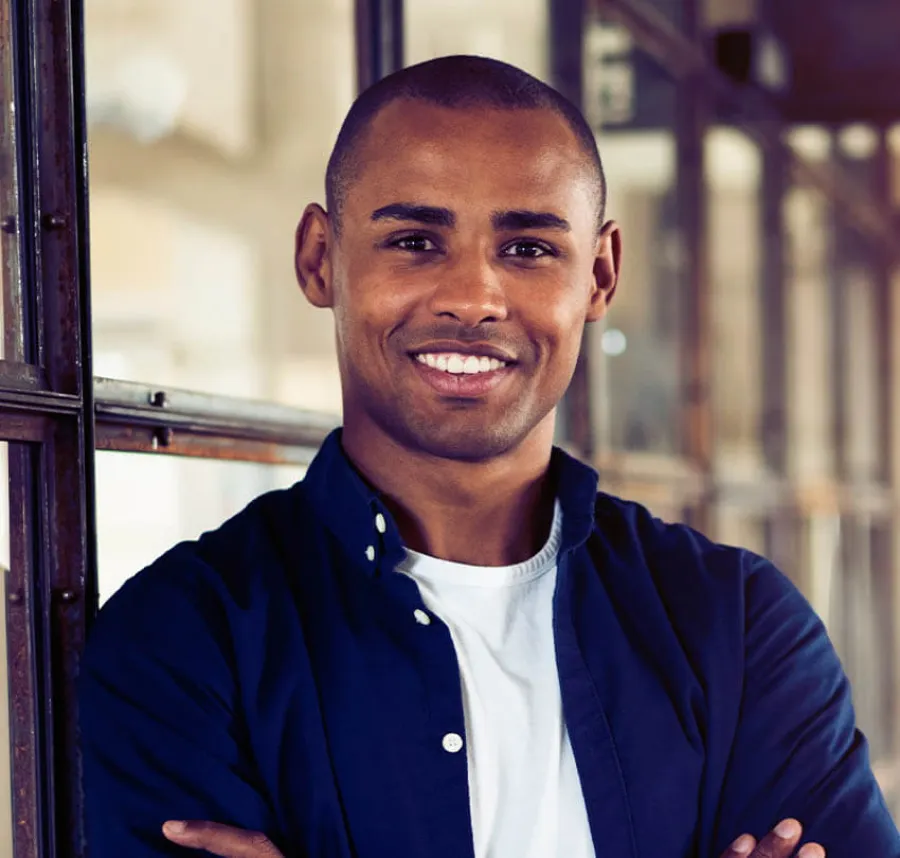 Confident young man with arms crossed smiling in casual blue shirt and white t-shirt indoors by glass window.