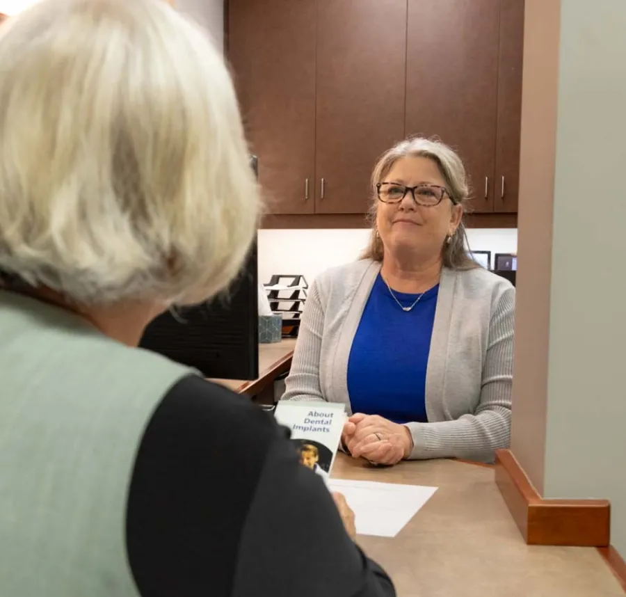 Patient consulting with a female receptionist at a beige and brown medical office desk.