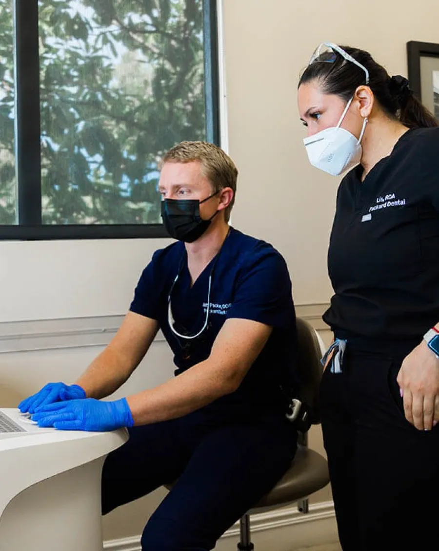 Two dental professionals wearing masks and gloves reviewing patient data on a computer in a dental office.