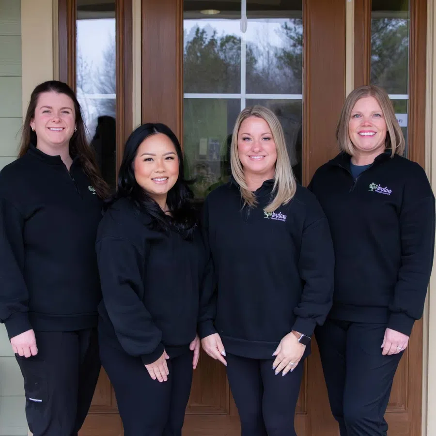 Four women in black matching sweatshirts standing in front of a wooden door smiling at camera