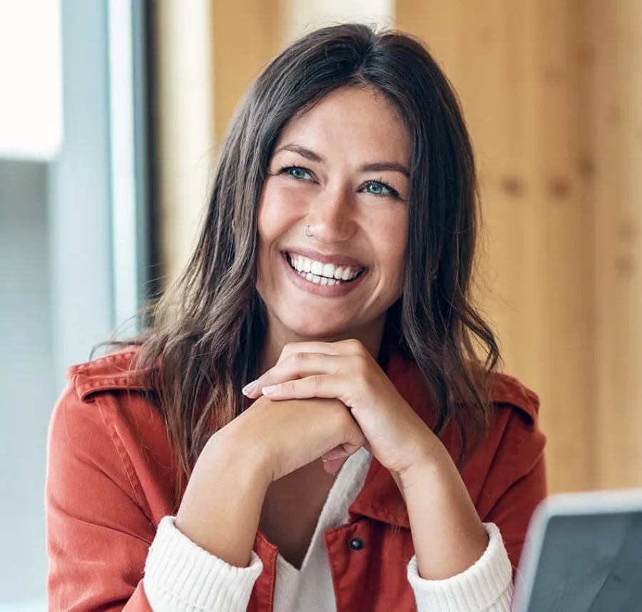 Smiling woman with long brown hair wearing a red jacket, sitting indoors with hands clasped near face.