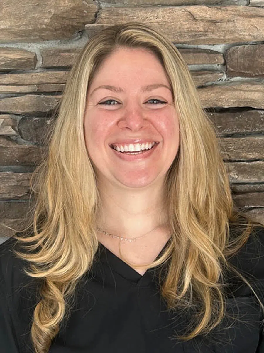 Smiling woman with long blonde hair wearing a black top standing in front of a stone wall background