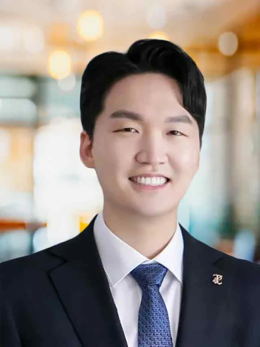 Young professional man smiling wearing a dark suit, white shirt, and blue patterned tie in a bright office.