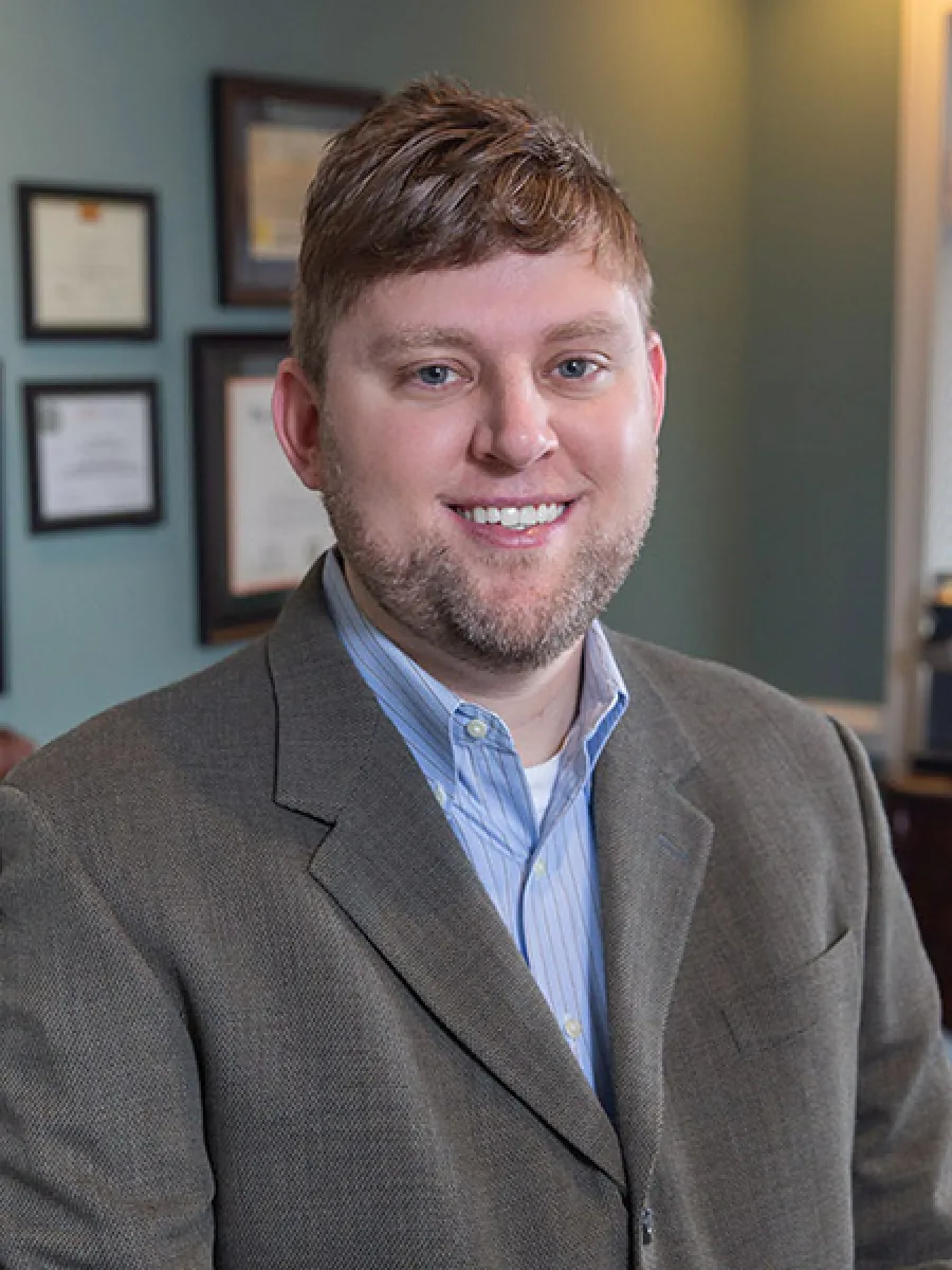 Smiling man in a brown suit jacket and blue striped shirt posing in an office with framed certificates on the wall.