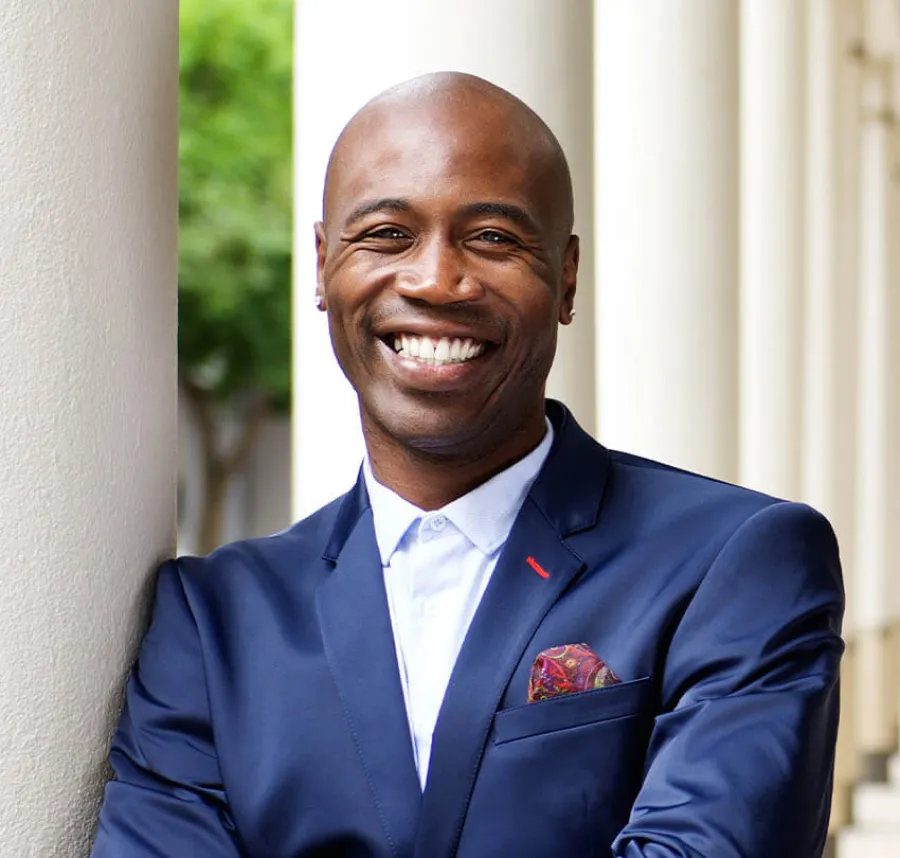 Smiling bald man in navy suit with patterned pocket square leans on white column outdoors with blurred background.