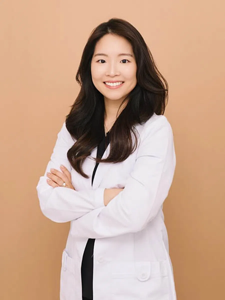 Smiling female doctor with long hair wearing a white lab coat standing with crossed arms on beige background.