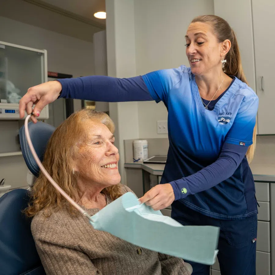 Dental hygienist smiling while placing a protective bib on an elderly woman patient in a clinic.