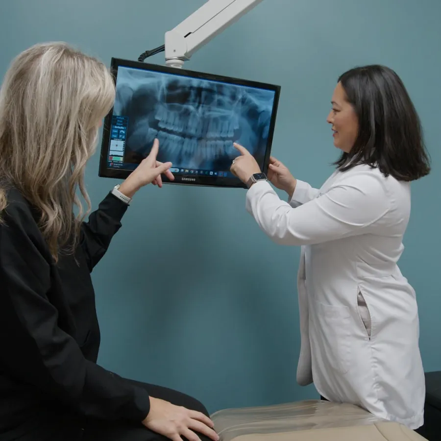 Female dentist and patient examining dental X-ray on monitor in dental office with blue wall background