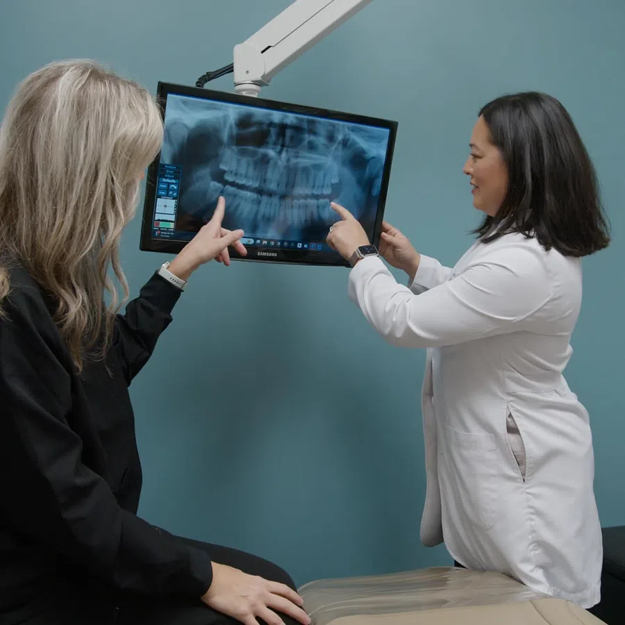 Female dentist and patient examining dental X-ray on monitor in dental office with blue wall background