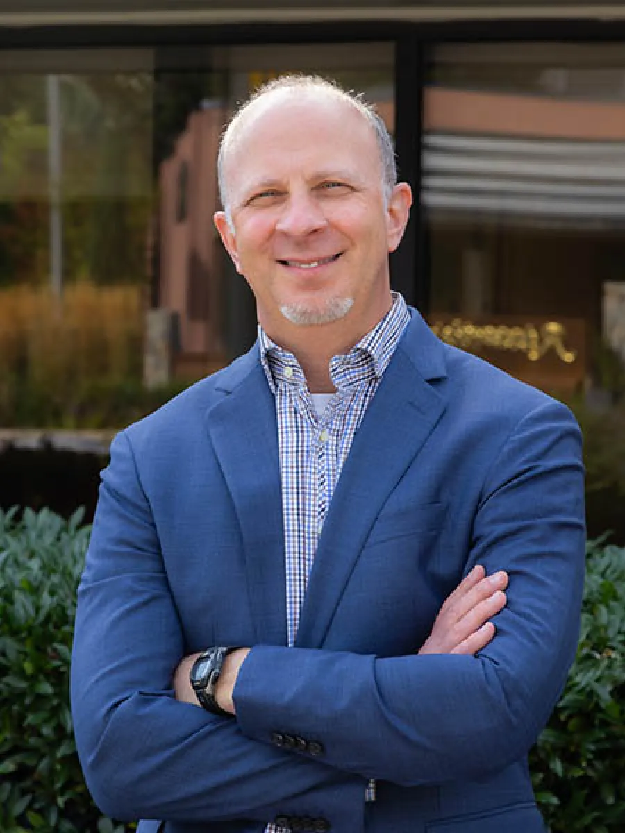 Smiling middle-aged man in a blue blazer and checkered shirt stands outdoors with arms crossed.