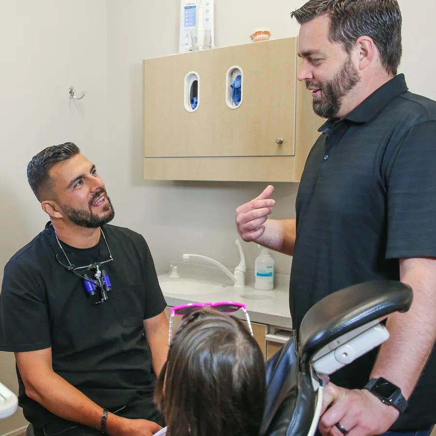 A dentist discussing with a patient while a child observes in a dental office setting.