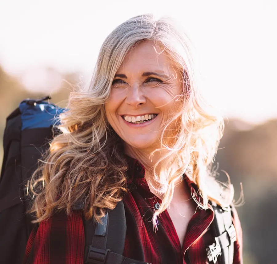 Smiling middle-aged woman with blonde hair wearing a red plaid shirt and hiking backpack outdoors in sunlight