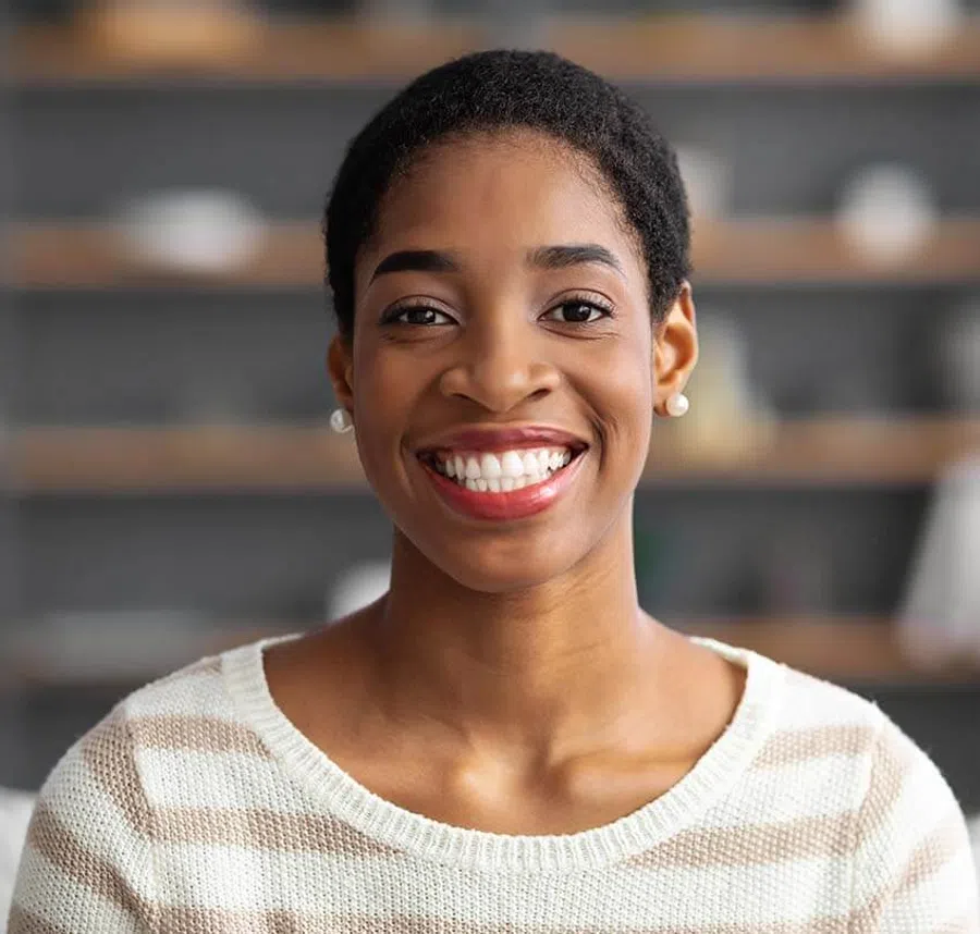 Smiling young woman with short curly hair wearing pearl earrings and a striped sweater indoors.