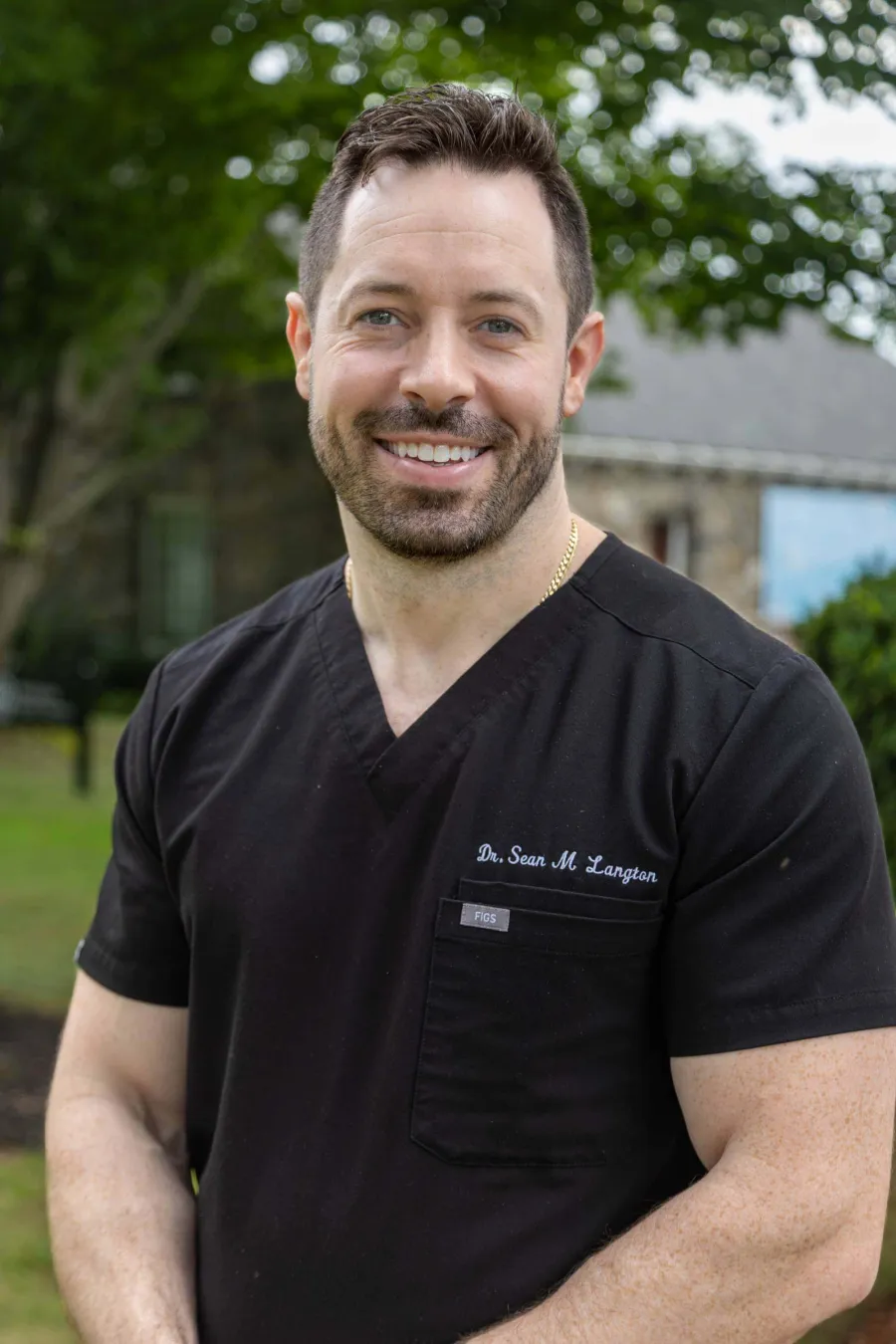Smiling male doctor wearing black scrubs standing outdoors with greenery and buildings in the background.