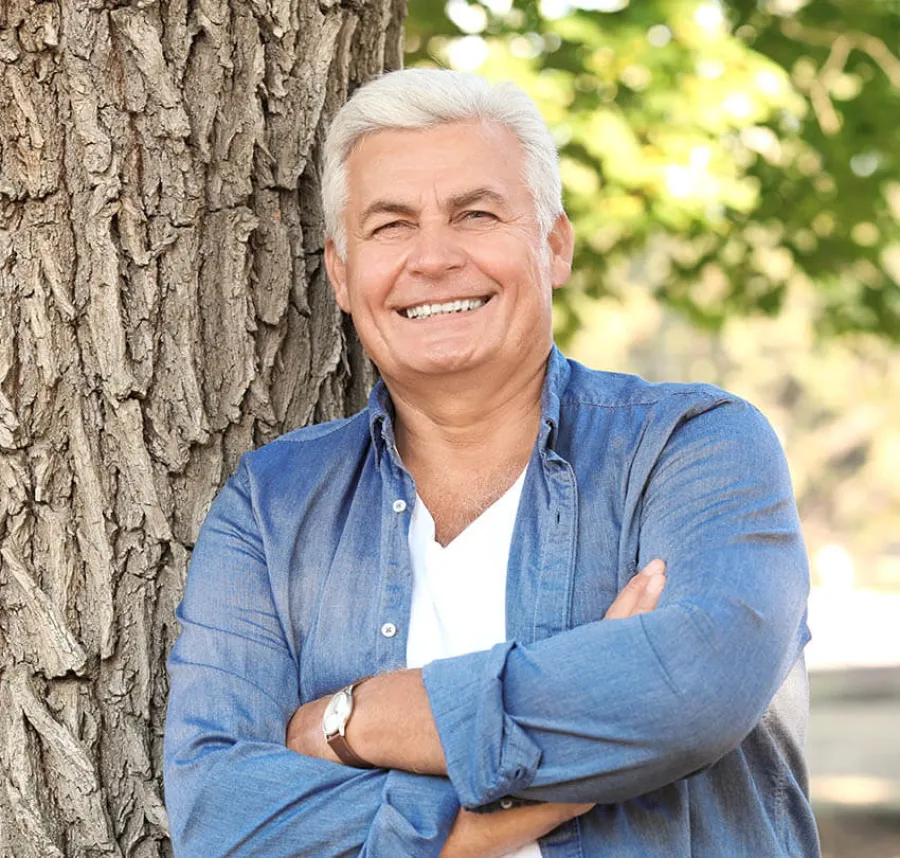 Smiling mature man with white hair in blue shirt standing outdoors, leaning against a tree with crossed arms.