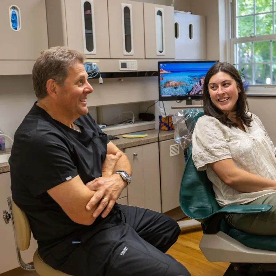 Dentist and patient smiling and talking in a bright dental office with equipment and monitors.