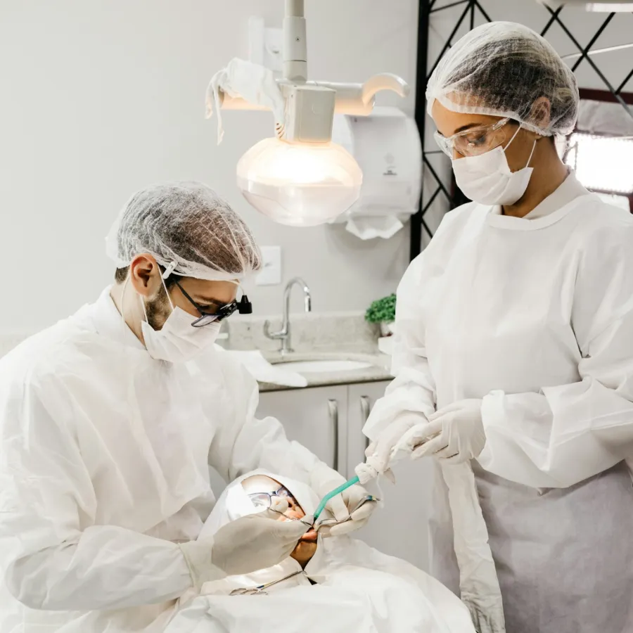 Dentist and assistant in protective gear performing dental procedure on patient under bright dental light.