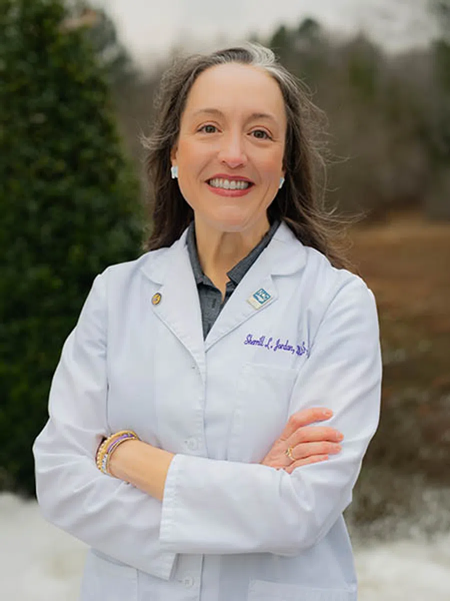 Smiling female doctor in white lab coat standing outdoors with arms crossed and blurred background.