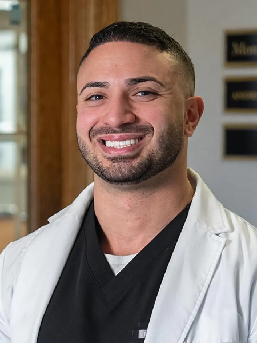 Smiling male healthcare professional wearing a white coat and black scrubs in an office setting