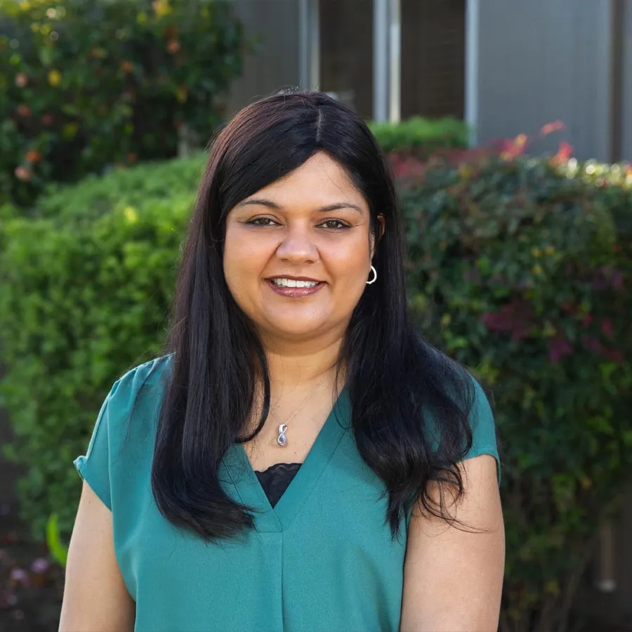 Portrait of a smiling woman with long black hair wearing a teal blouse standing outdoors with greenery in background