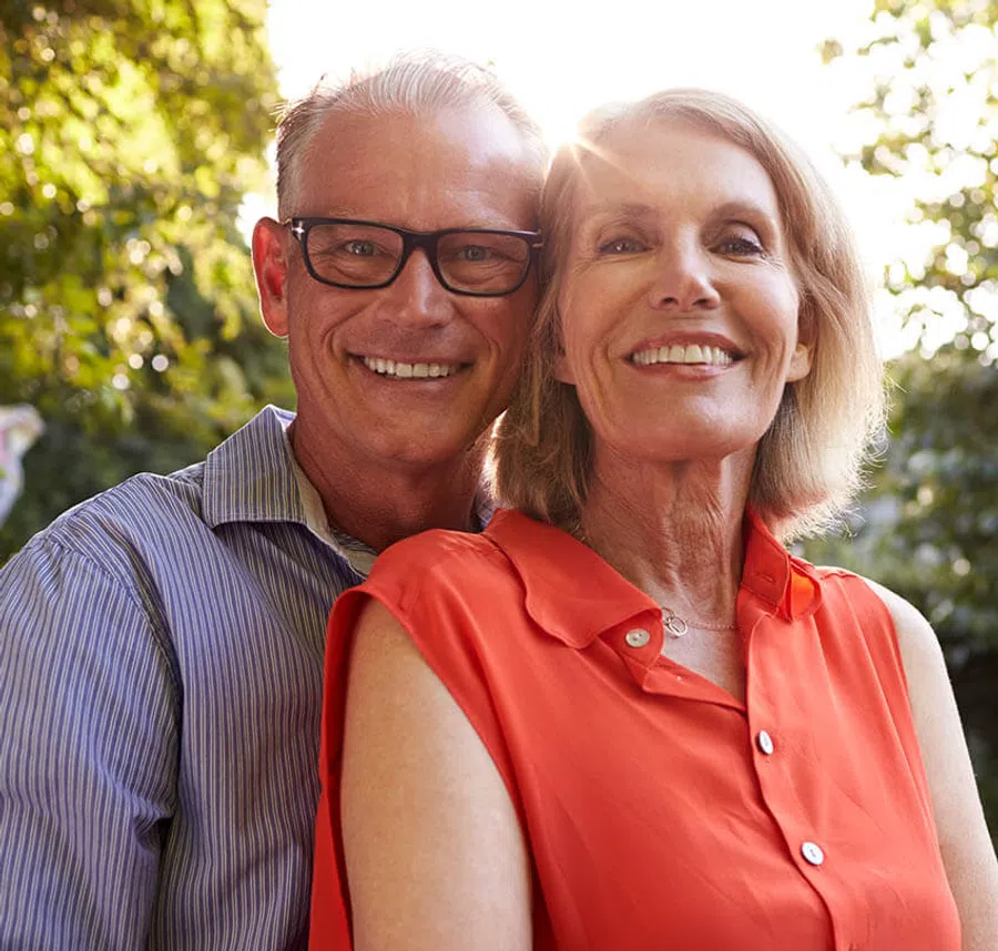 Smiling mature couple outdoors with sunlit greenery, man in glasses and striped shirt, woman in orange blouse