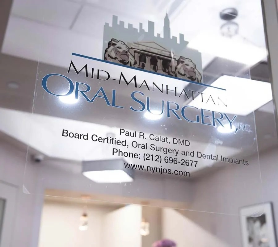 Glass door sign for Mid-Manhattan Oral Surgery with contact details and website in clinic office.