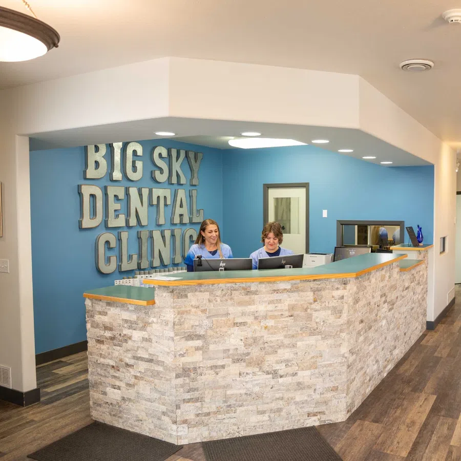 Reception area at Big Sky Dental Clinic with two staff members working behind stone front desk and blue accent wall.