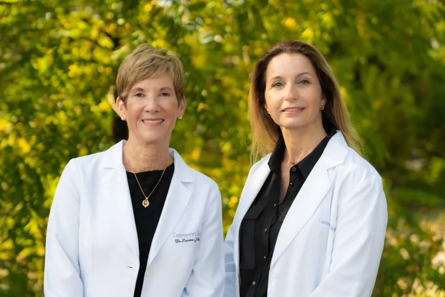 Two female doctors in white coats smiling outdoors with green trees in the background.