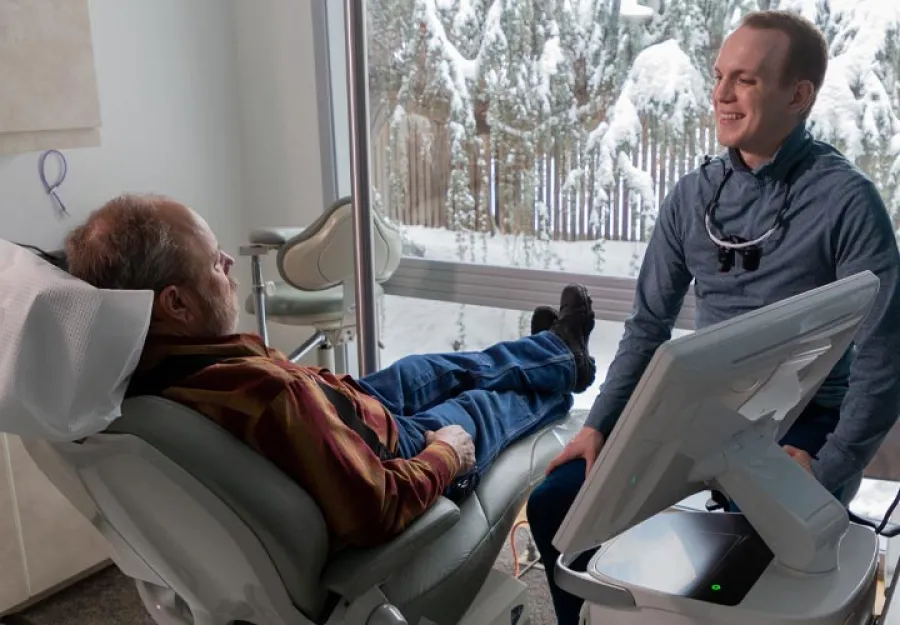 Dentist consulting with elderly male patient reclining in dental chair overlooking snowy outdoor scene.