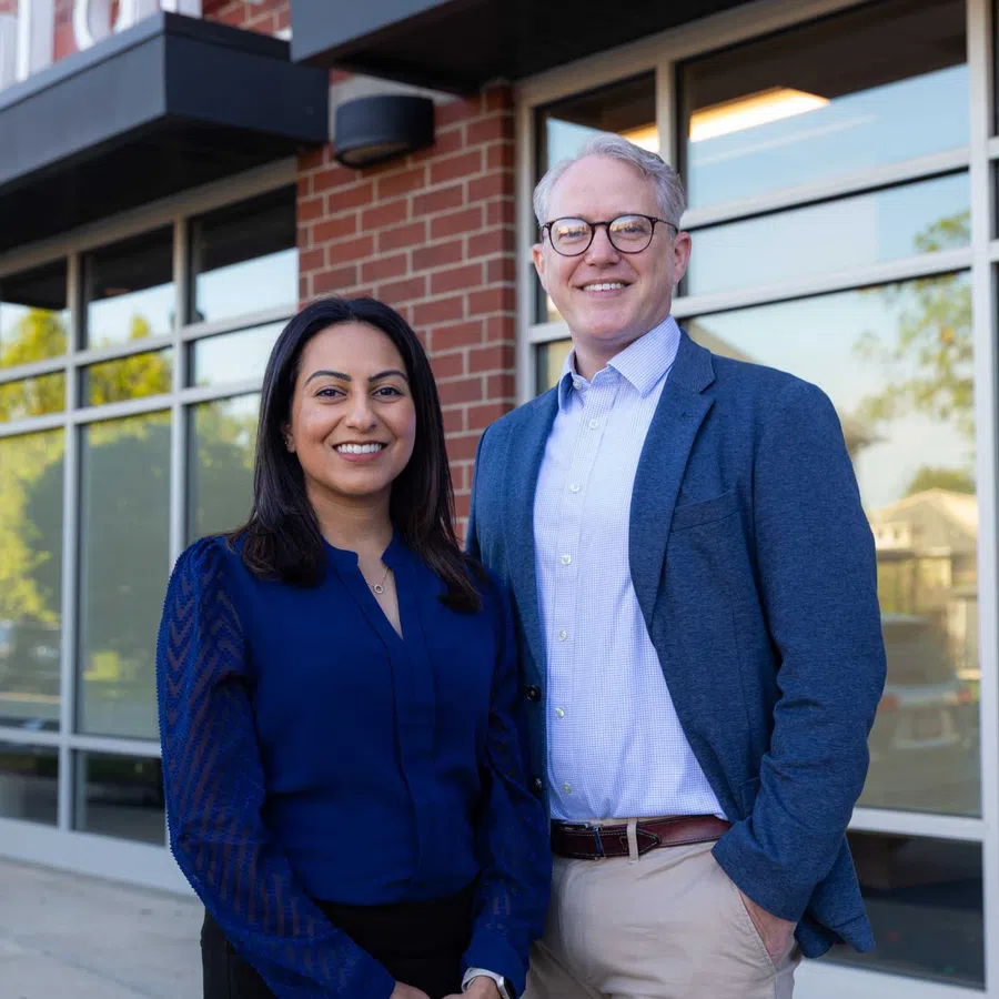 Two dental professionals smiling outside a modern dental office with large glass windows and brick exterior.