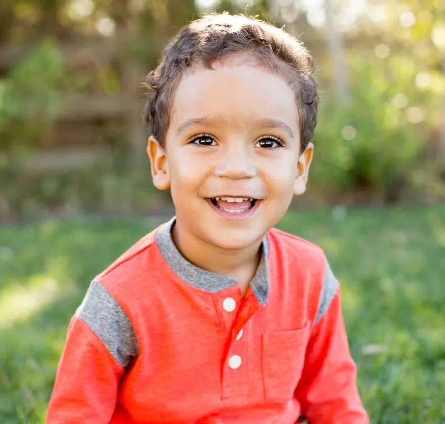 Smiling young boy in an orange and gray shirt sitting outdoors on green grass in soft sunlight.