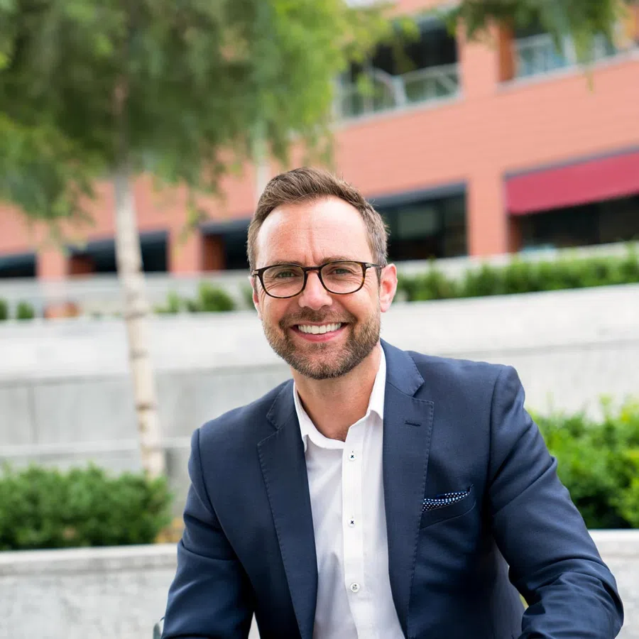 Smiling man in glasses and business suit sitting outdoors with blurred urban background and greenery.