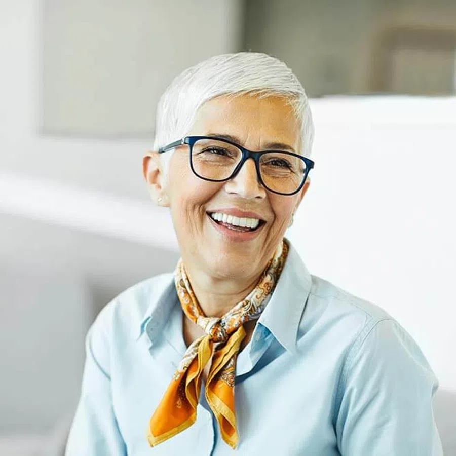 Smiling elderly woman with short white hair, wearing glasses, a blue shirt, and an orange patterned scarf.
